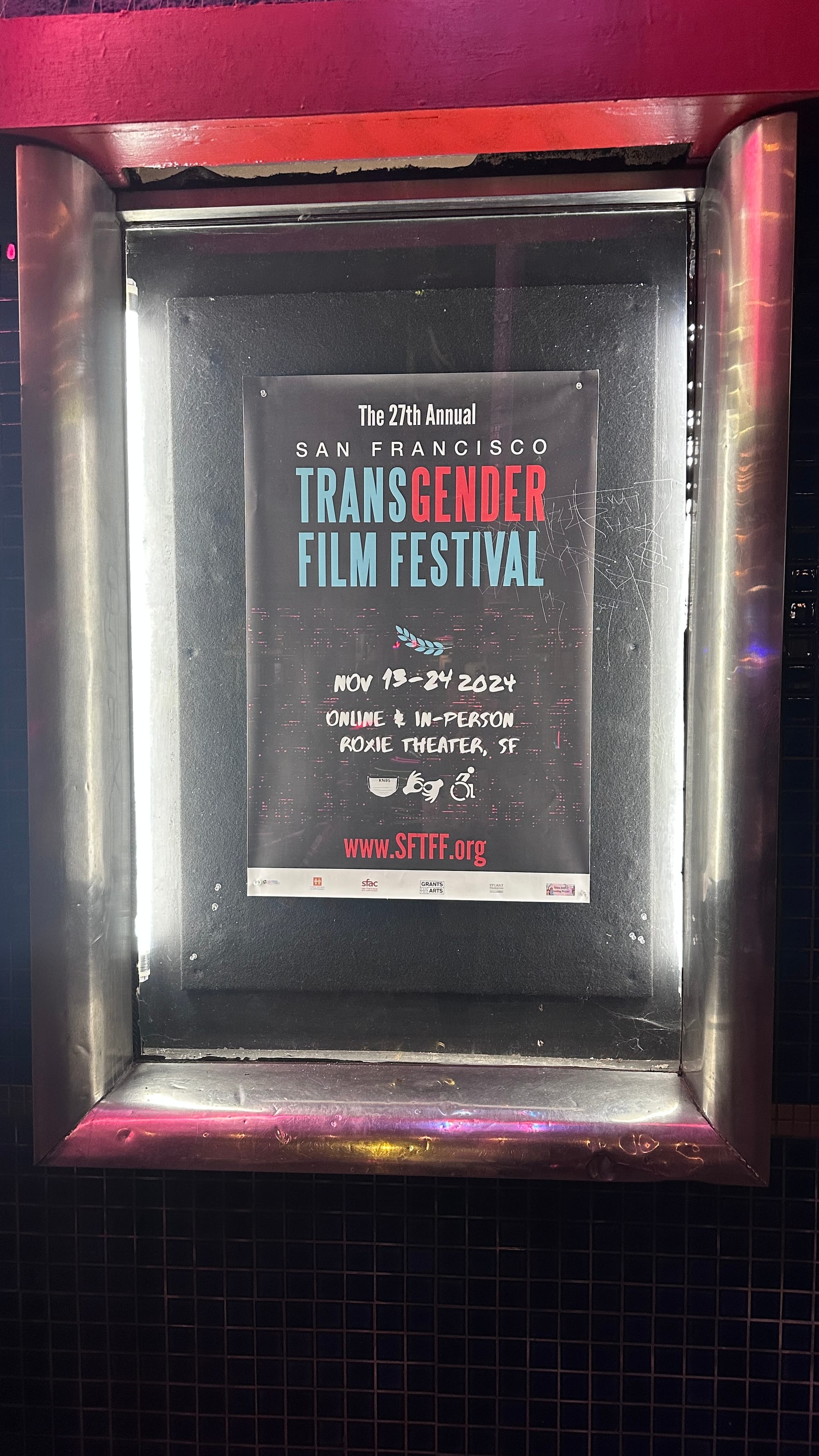 A framed poster for "The 27th Annual San Francisco Transgender Film Festival" displayed on an exterior wall. The poster highlights the event dates, "Nov 13-24 2024," and mentions the online and in-person venue at the Roxie Theater in San Francisco. The festival's website, "www.SFTFF.org," is prominently featured at the bottom, with additional logos and credits below.