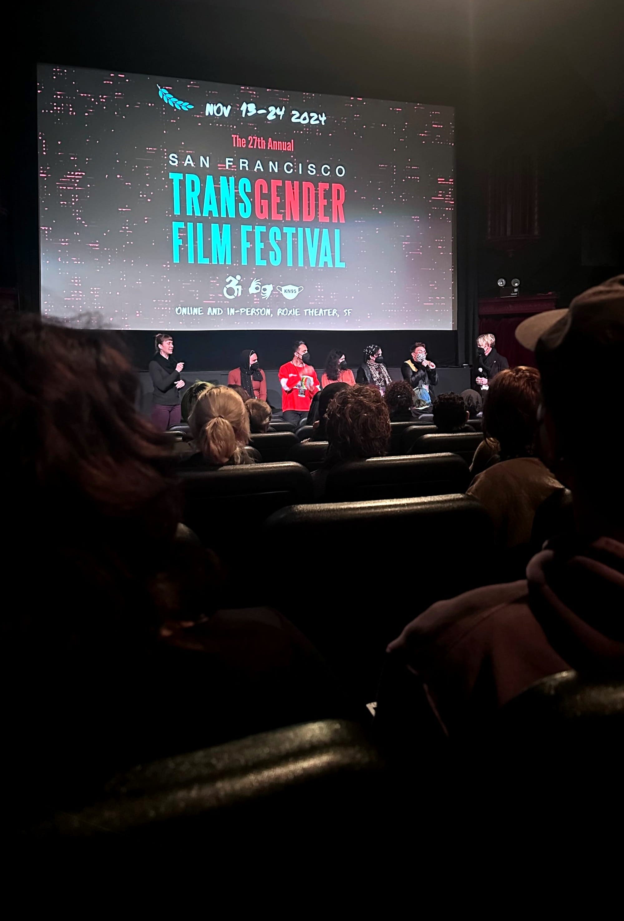 A dimly lit theater with rows of audience members facing a large screen that displays the title "The 27th Annual San Francisco Transgender Film Festival." The screen includes the dates "Nov 13-24 2024" and mentions that the festival is taking place both online and in-person at the Roxie Theater in San Francisco. Onstage, several people are participating in a panel discussion, with one person speaking into a microphone.